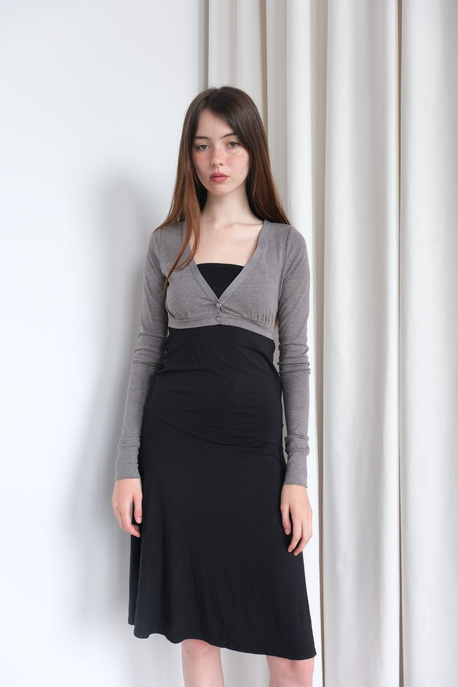 Woman wearing a black dress with a gray cardigan against a white curtain background