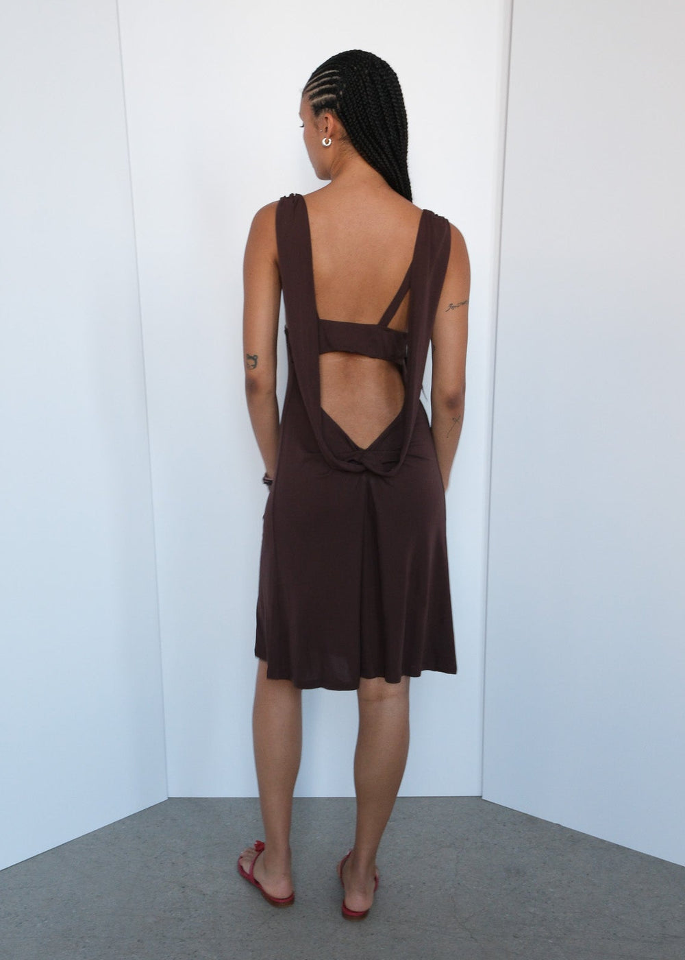 Woman wearing a brown dress with cut-out back design against a white wall.