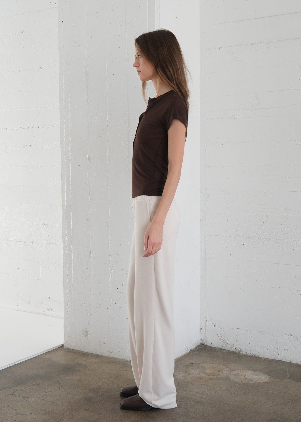 Woman wearing a brown top and white pants standing against a white wall.