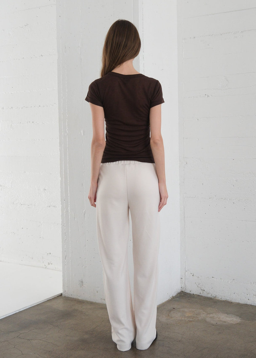 Person wearing a brown top and white pants standing against a white brick wall.