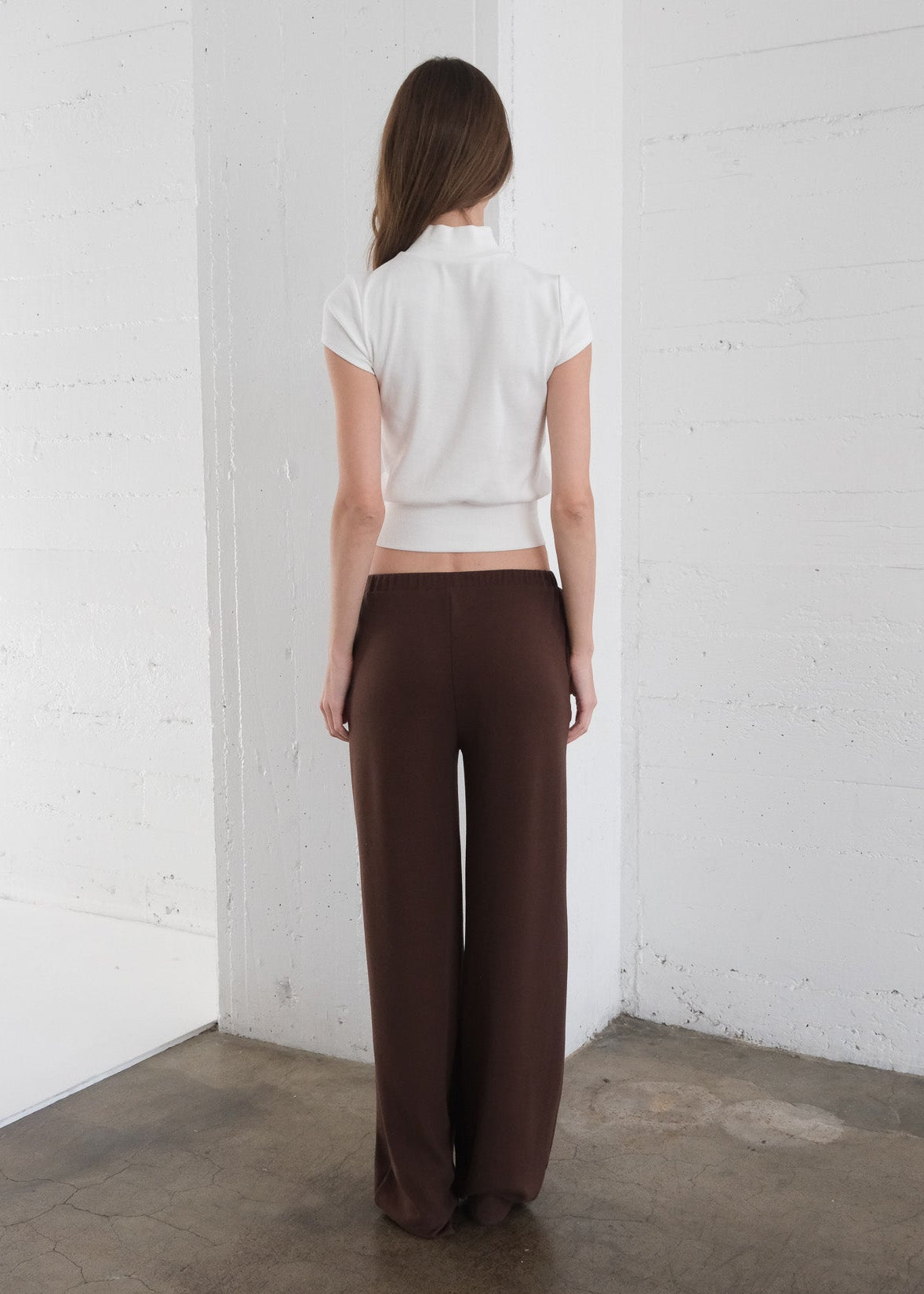 Woman wearing a white top and brown pants against a white brick wall.