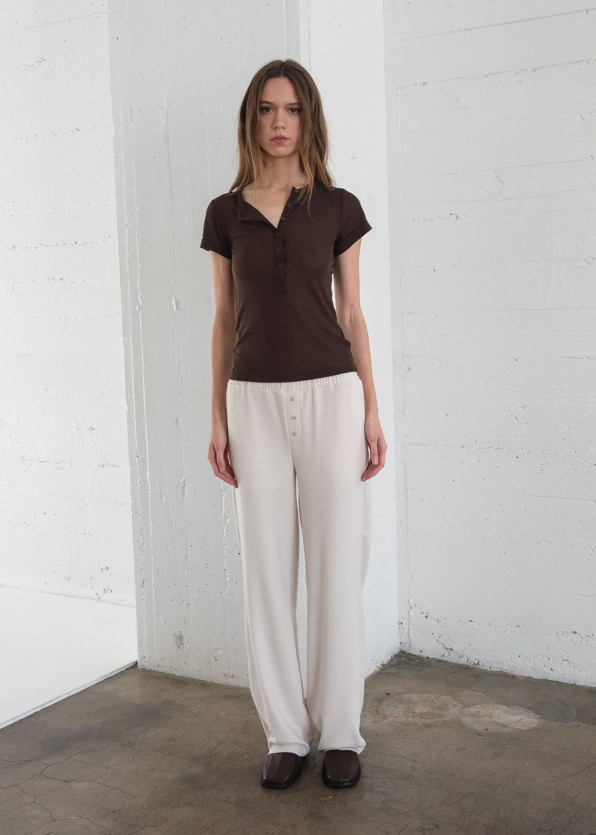 Woman wearing a brown shirt and white pants against a white wall.